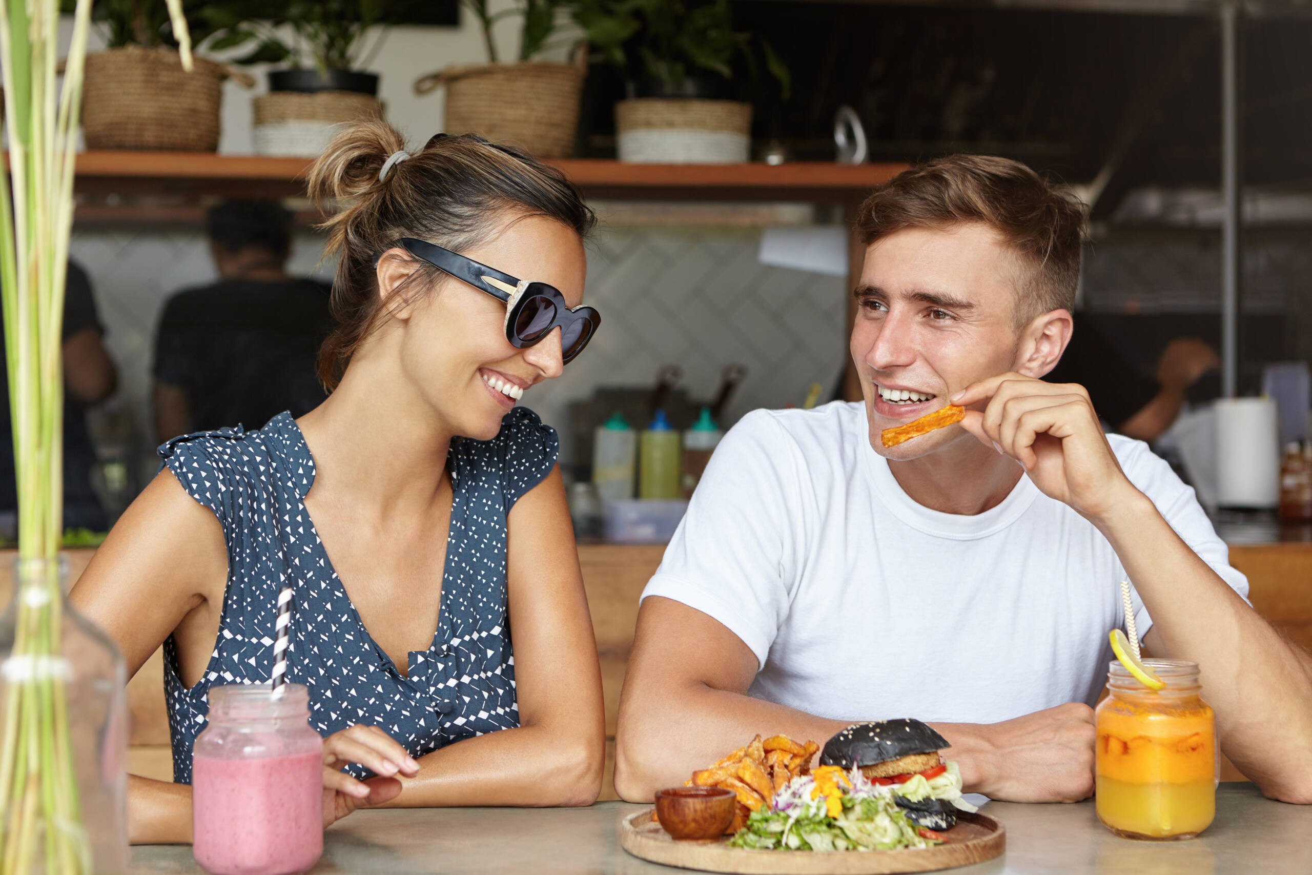 Love and friendship. Happy couple eating burger with French fries and having fresh drinks during date at cozy cafeteria. Cute woman in trendy sunglasses listening to her boyfriend’s jokes and laughing